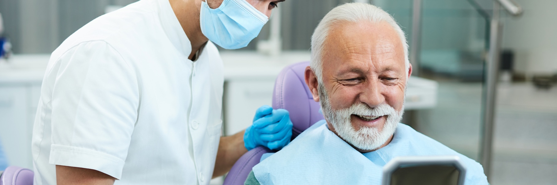 Smiling older male patient in dental chair reviewing results with masked dentist nearby.